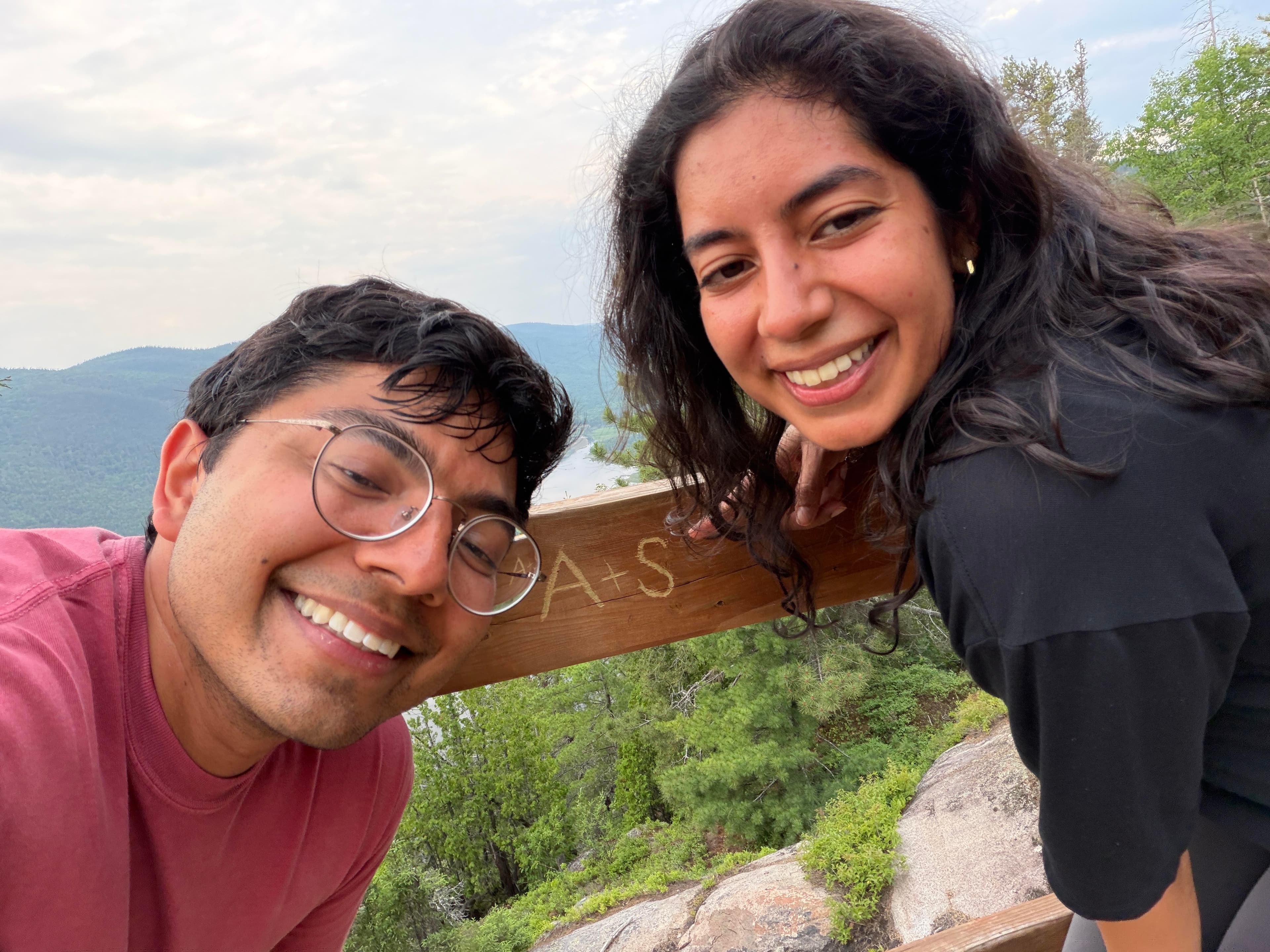 Anahita and Snehan smiling together in the mountains beside a wooden rail marked A plus S
